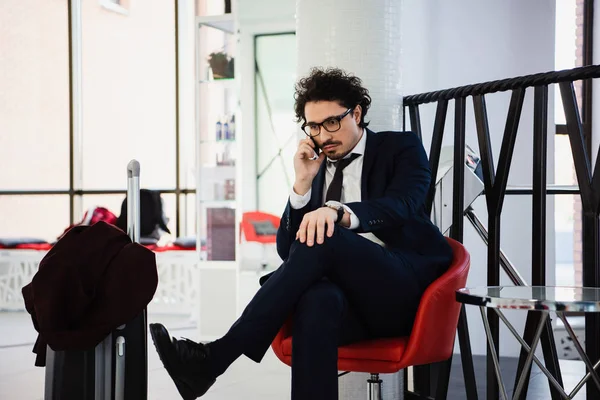 Serious businessman talking on smartphone while waiting in hotel lobby with suitcase — Stock Photo