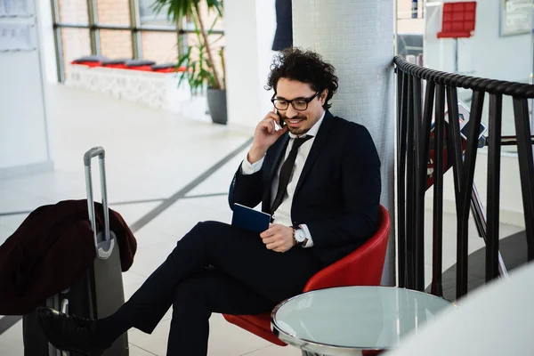 Homem de negócios sorrindo falando no smartphone e olhando para o passaporte enquanto espera no lobby do hotel com mala — Fotografia de Stock