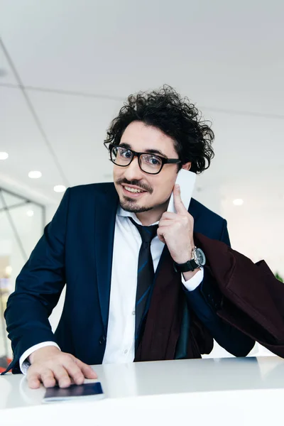 Uomo d'affari sorridente con passaporto che parla su smartphone mentre effettua il check-in in hotel — Stock Photo