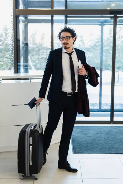 Бизнесмен со смартфоном, паспортом и дорожной сумкой, стоящий на стойке регистрации в отеле — стоковое фото