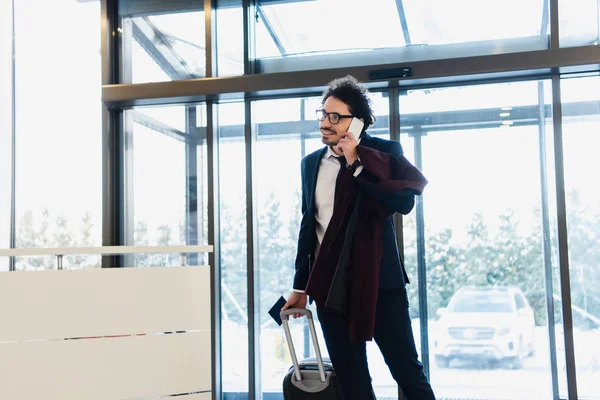 Sonriente hombre de negocios con pasaporte y equipaje hablando en smartphone en el vestíbulo del hotel — Stock Photo