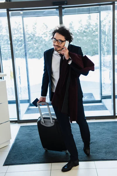 Smiling businessman with passport and travel bag talking on smartphone in hotel hall — Stock Photo