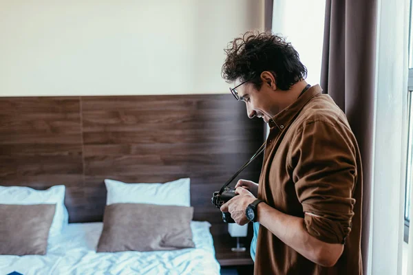 Männlicher Fotograf mit Brille blickt auf Fotokamera im Hotelzimmer — Stockfoto