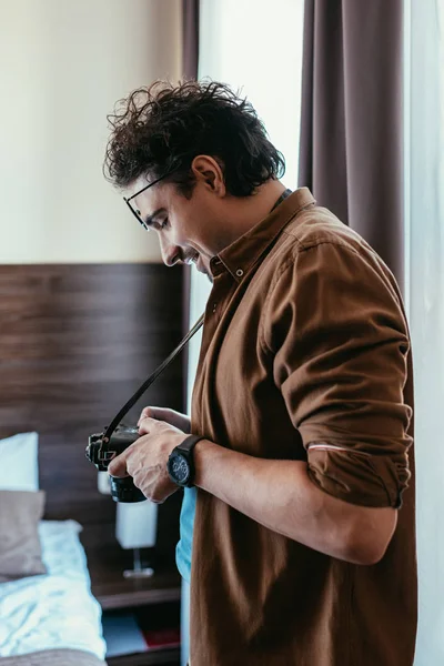 Lächelnder Fotograf mit Brille blickt auf Fotokamera im Hotelzimmer — Stockfoto