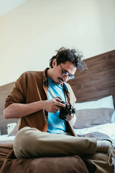 Homem adulto em óculos olhando para a câmera fotográfica e sentado na cama no quarto do hotel — Fotografia de Stock