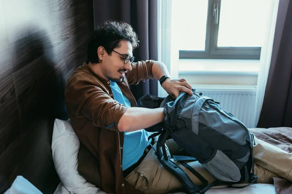 Красивий чоловік мандрівник бере щось з рюкзака на ліжку в готельному номері — стокове фото
