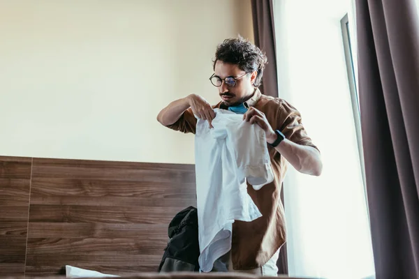 Turista masculino en anteojos poniendo camisa en mochila en habitación de hotel - foto de stock