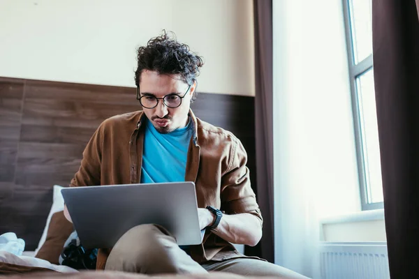 Confused man using laptop on bed in hotel room — Stock Photo