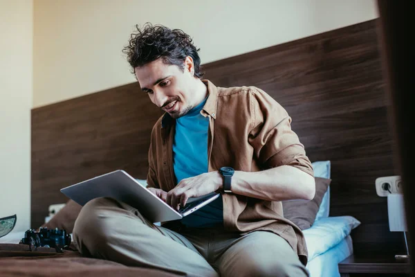 Smiling traveler using laptop in hotel room with photo camera on bed — Stock Photo