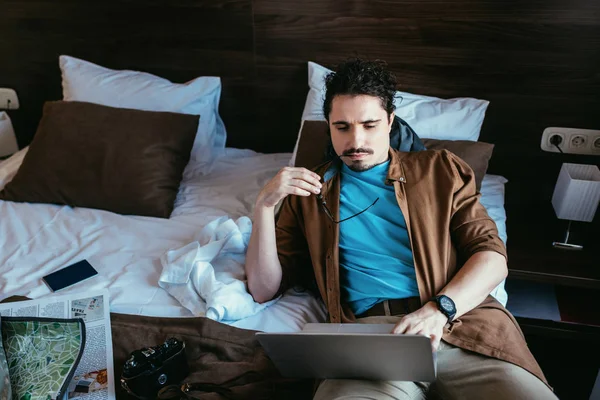 Pensive traveler using laptop in hotel room with newspaper, map, smartphone and photo camera on bed — Stock Photo