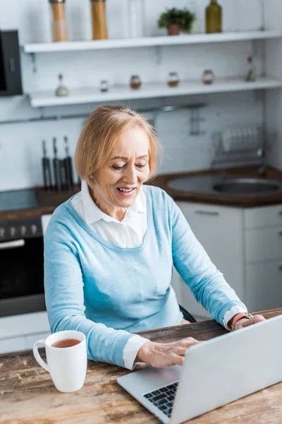 Sorrindo mulher idosa sentada à mesa com xícara de chá e usando laptop na cozinha — Fotografia de Stock