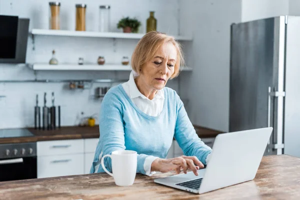 Donna anziana seduta a tavola con tazza di caffè e utilizzando il computer portatile in cucina — Foto stock