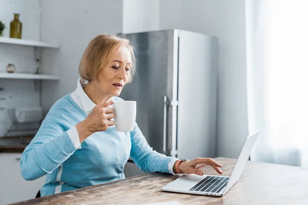 Mujer mayor sentada en la mesa, bebiendo café y utilizando el ordenador portátil en la cocina con espacio para copias - foto de stock