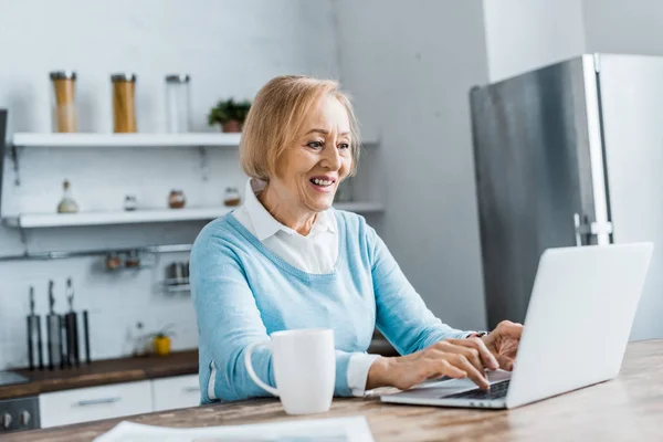 Donna anziana sorridente seduta a tavola con tazza di caffè e utilizzando il computer portatile in cucina — Foto stock