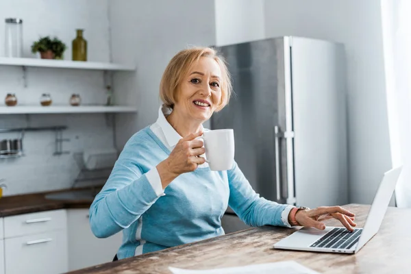 Femme âgée souriante assise à table, regardant la caméra, buvant du café et utilisant un ordinateur portable dans la cuisine — Photo de stock