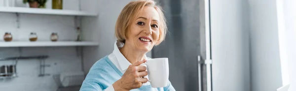 Sonriente mujer mayor sosteniendo una taza de café y mirando a la cámara en casa - foto de stock
