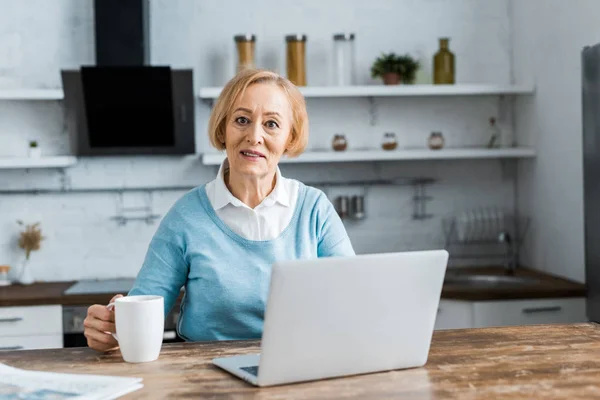 Femme âgée assise à table avec une tasse de café, regardant la caméra et utilisant un ordinateur portable dans la cuisine — Photo de stock