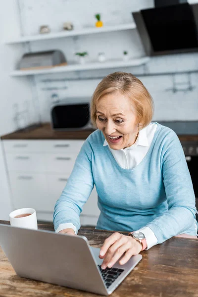 Mulher sênior animado sentado à mesa com xícara de chá e usando laptop na cozinha — Fotografia de Stock