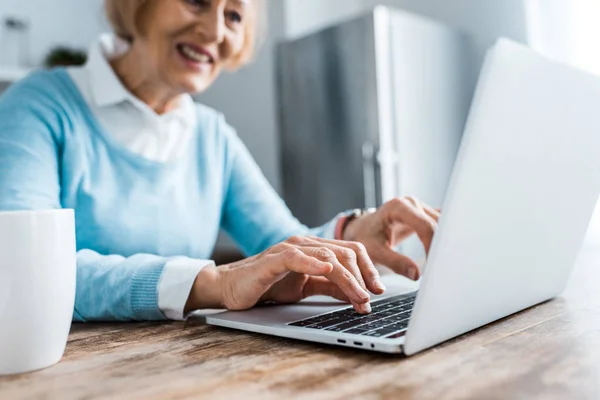 Foco seletivo de sorrir mulher idosa digitando no laptop em casa — Fotografia de Stock