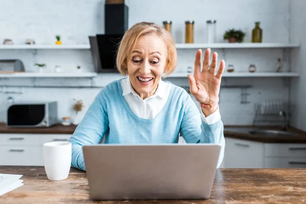 Felice donna anziana salutando con mano, utilizzando il computer portatile e avendo video chat in cucina — Foto stock