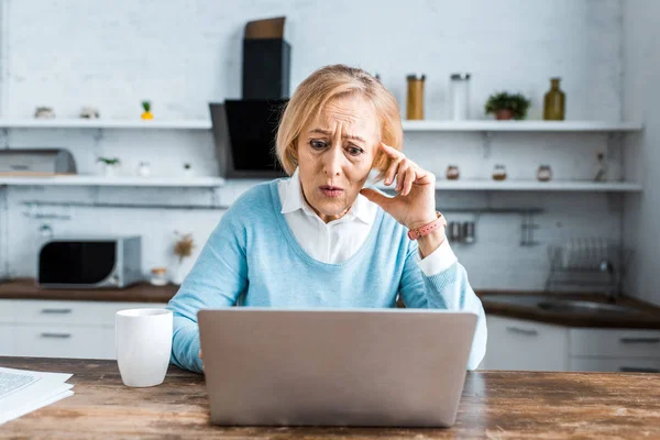 Shocked senior woman touching face, using laptop and having video chat in kitchen — Stock Photo