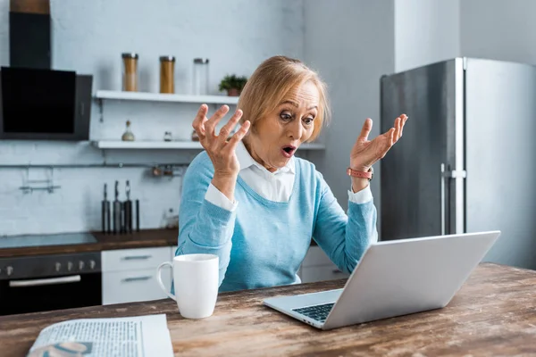 Здивована старша жінка жестикулює руками, використовуючи ноутбук і маючи відеочат на кухні — стокове фото