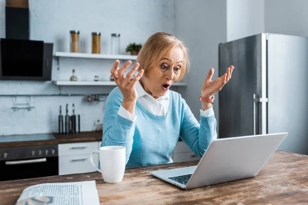 Überraschte Seniorin gestikulierte mit Händen, benutzte Laptop und führte Videochat in Küche — Stockfoto