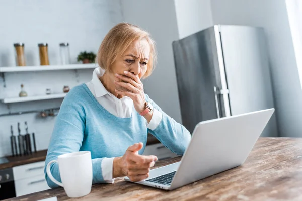 Sconvolto donna anziana che copre la bocca con la mano, utilizzando il computer portatile e avendo video chat in cucina — Foto stock