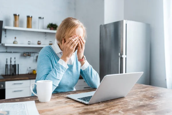 Стресс старшая женщина сидит за столом и покрывает лицо во время видеозвонка на ноутбук на кухне — стоковое фото