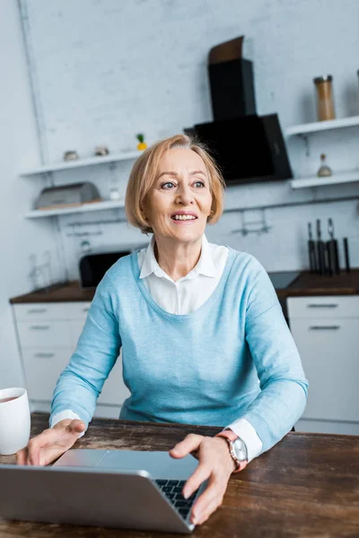 Улыбающаяся пожилая женщина в повседневной одежде сидит за столом с ноутбуком на кухне — стоковое фото