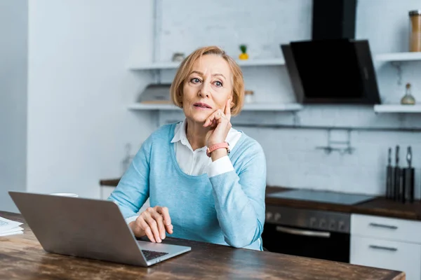 Pensive senior woman sitting at table with laptop and propping chin in kitchen — Stock Photo