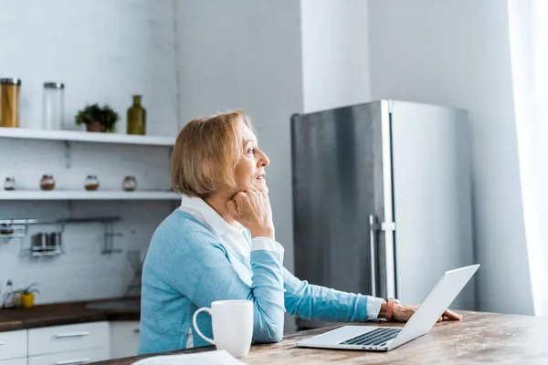 Pensive senior woman sitting at table with cup of coffee and laptop in kitchen — Stock Photo