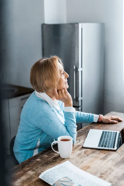 Pensive senior woman sitting at table with cup of coffee and laptop in kitchen — Stock Photo