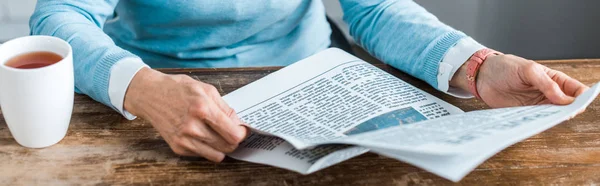 Vista cortada da mulher sênior sentada à mesa com xícara de chá e jornal de leitura em casa — Fotografia de Stock