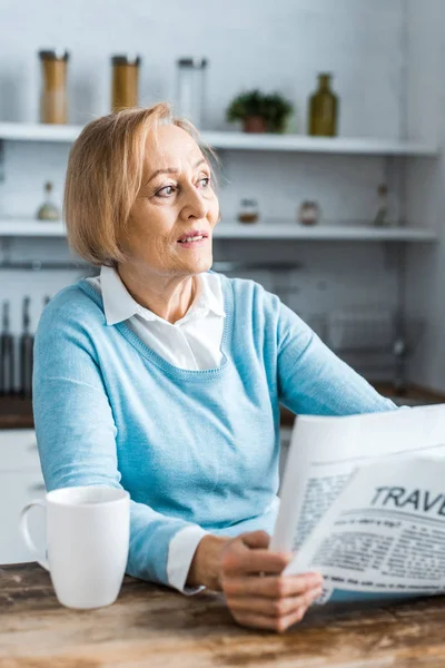 Senior woman sitting at table with cup of coffee and holding travel newspaper at home — Stock Photo