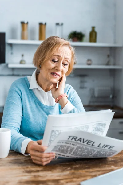 Senior woman sitting, touching face and reading travel newspaper at home — Stock Photo