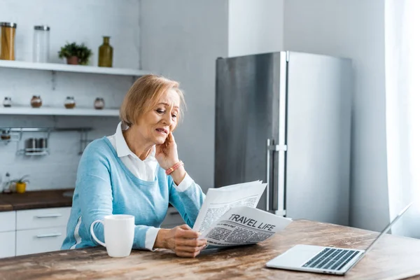 Mujer mayor sentada en la mesa con una taza de café y un portátil mientras lee el periódico en la cocina - foto de stock