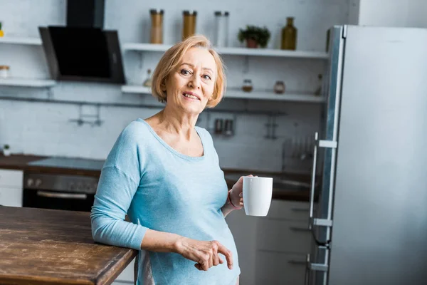 Lächelnde Seniorin mit Tasse Kaffee blickt in Küche in die Kamera — Stockfoto