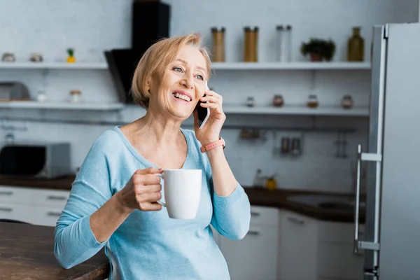 Femme âgée souriante avec tasse de café parler sur smartphone à la maison — Photo de stock
