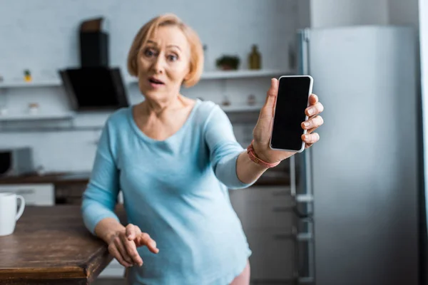 Seniorin blickt in Kamera und präsentiert Smartphone mit leerem Bildschirm zu Hause — Stockfoto
