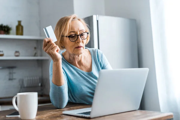 Donna anziana in occhiali in possesso di carta di credito e utilizzando il computer portatile durante lo shopping online a casa — Foto stock