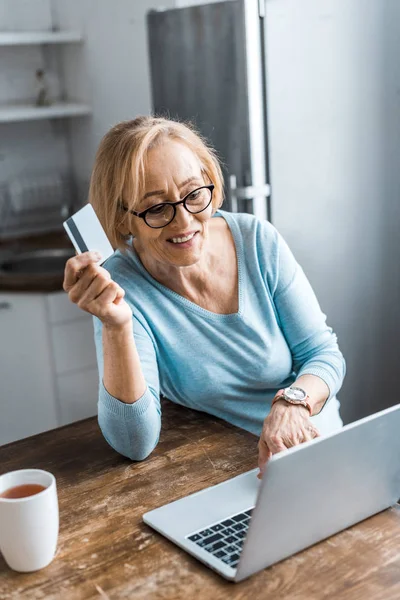 Sonriente mujer mayor sosteniendo la tarjeta de crédito y el uso de la computadora portátil mientras hace compras en línea en casa - foto de stock