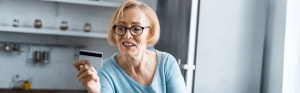 Femme âgée souriante dans des lunettes tenant la carte de crédit à la maison avec espace de copie — Photo de stock