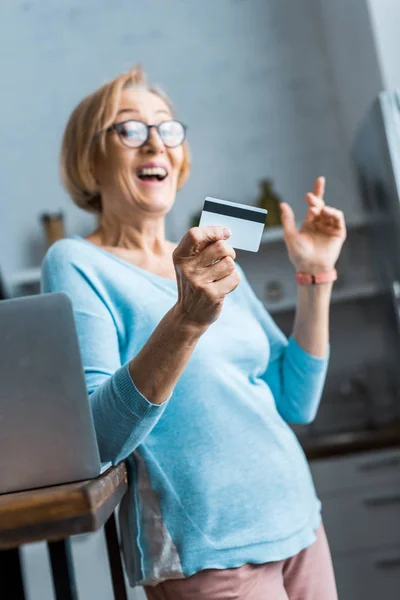 Excitée femme âgée dans des lunettes tenant carte de crédit près d'un ordinateur portable à la maison — Photo de stock