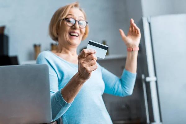 Mujer mayor excitada en gafas gestos con la mano y la celebración de la tarjeta de crédito cerca de la computadora portátil en casa - foto de stock