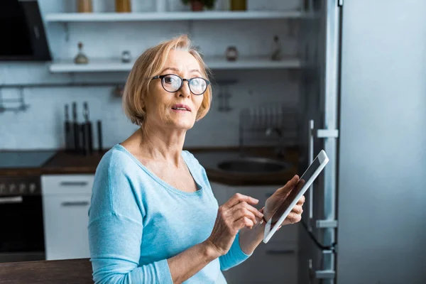 Mulher sênior em óculos olhando para a câmera e usando tablet digital em casa — Fotografia de Stock