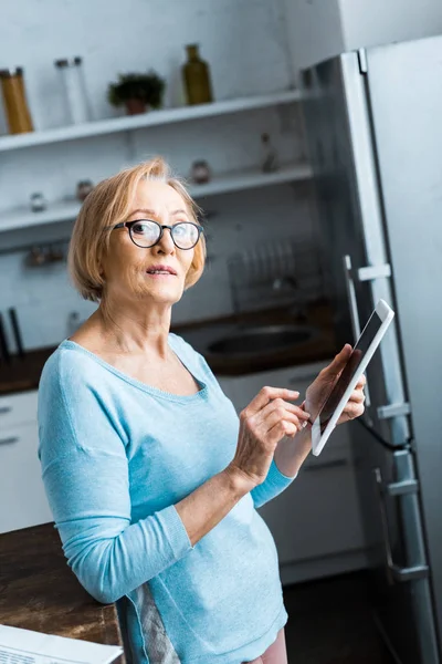 Seniorin mit Brille blickt auf Kamera und nutzt zu Hause digitales Tablet — Stockfoto