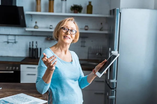 Donna anziana sorridente in bicchieri utilizzando tablet digitale e gesticolando con mano in cucina — Foto stock