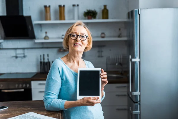 Senior woman in glasses looking at camera and presenting digital tablet with blank screen in kitchen — Stock Photo