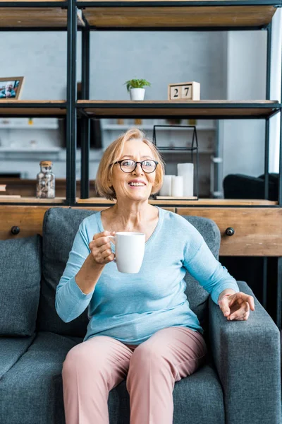 Улыбающаяся пожилая женщина в очках, сидящая на диване с чашкой кофе в гостиной — стоковое фото